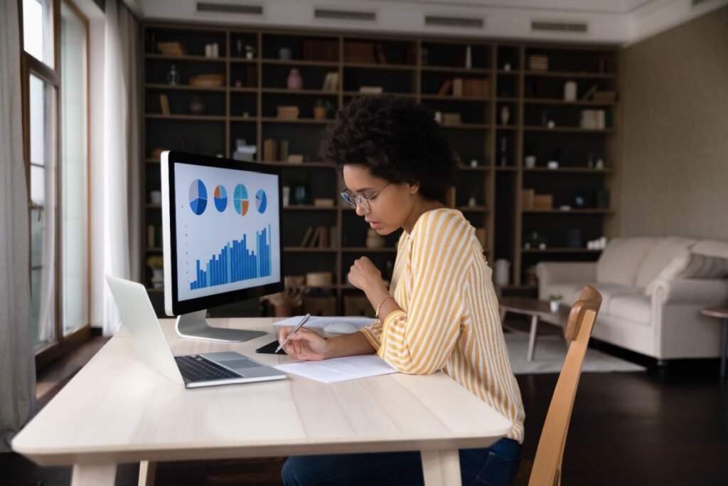 Mulher analisando gráficos financeiros no computador enquanto faz anotações