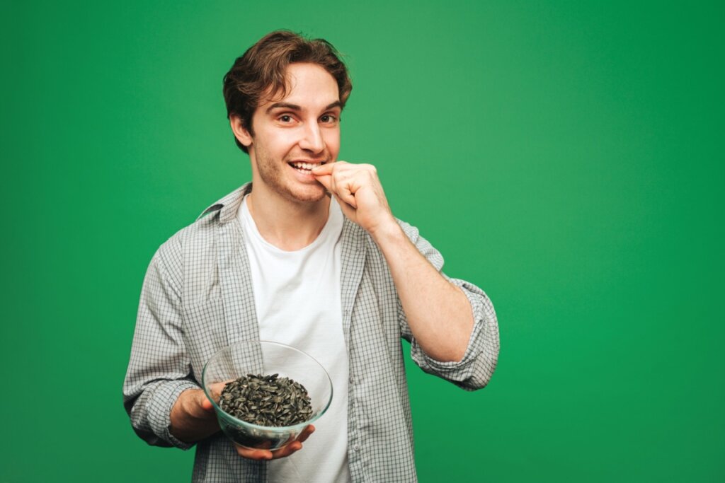 Homem usando uma camisa quadriculada por cima de uma branca, parado em frente a um fundo verde-escuro, e segurando um recipiente com sementes de girassol 