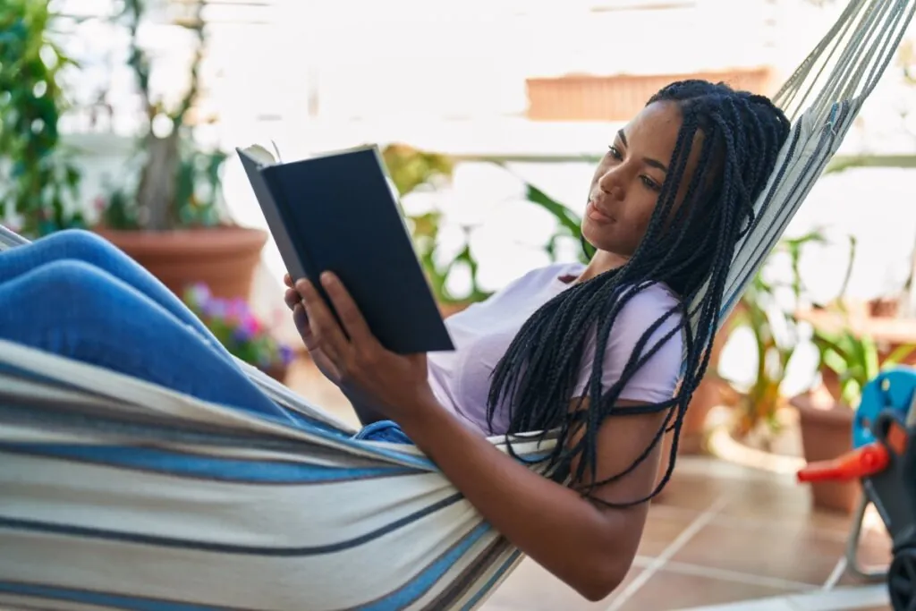Jovem com o cabelo longo, trançado, usando uma camiseta roxa deitada em rede listrada no quintal com um livro na mão