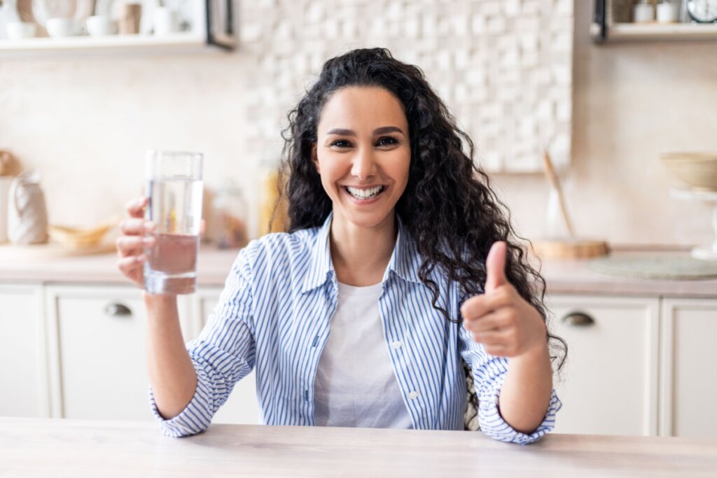 Mulher com cabelo longo cacheado e usando camiseta azul e camisa de botões listrada em azul e branco com um copo de água em uma mão e fazendo sinal positivo com a outra e sorrindo