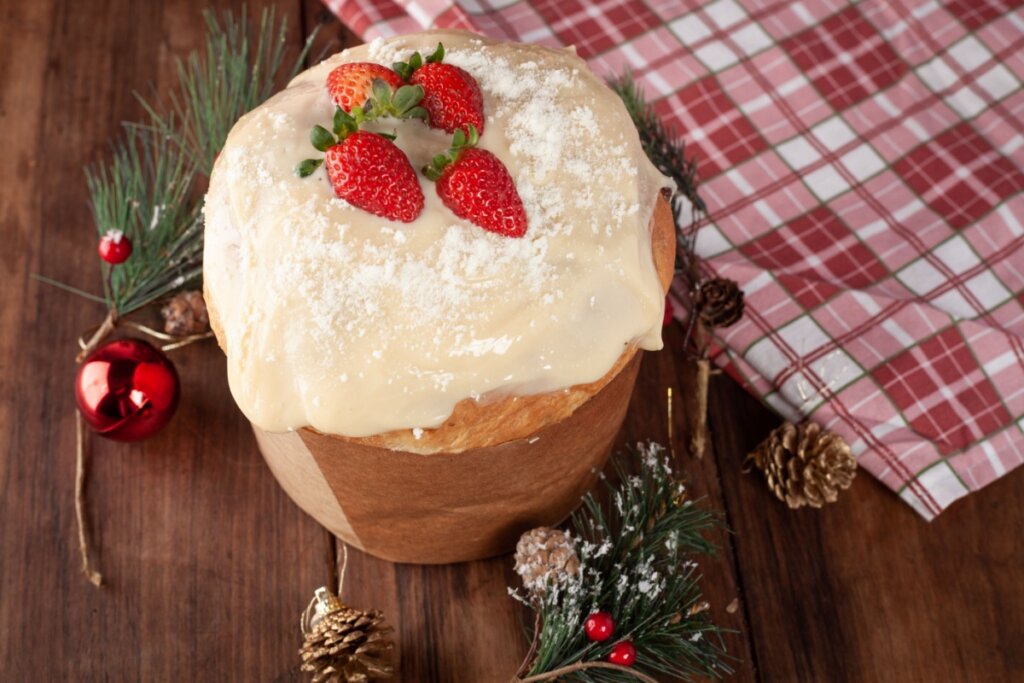Panetone coberto com creme branco e decorado com morangos inteiros sobre uma mesa de madeira com enfeites natalinos