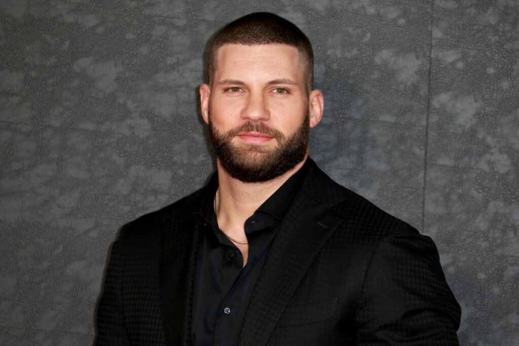 Homem usando blusa preta, parado em frente a um fundo cinza, e com o corte buzz cut e barba cheia modelada