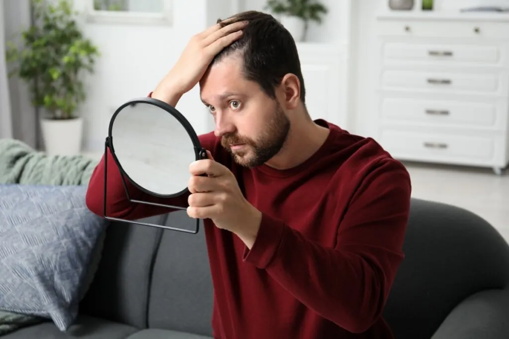 Homem com cabelo curto e barba usando blusa de manga longa vinho passando a mão no cabelo com uma mão e a outra segurando um espelho