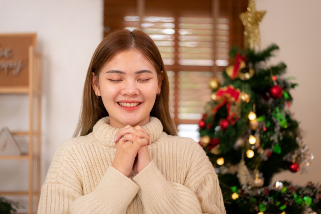 Jovem asiática com o cabelo solto, usando suéter bege rezando com árvores de Natal ao fundo