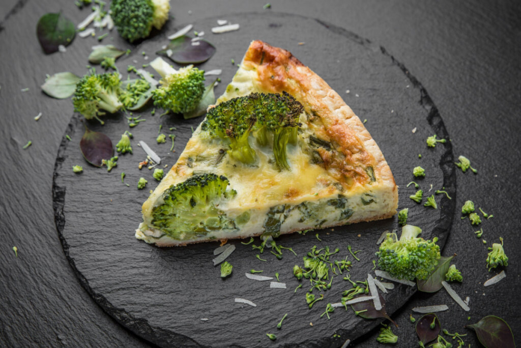 Pedaço de quiche de brócolis em cima de uma tábua de madeira preta com floretes de brócolis ao lado