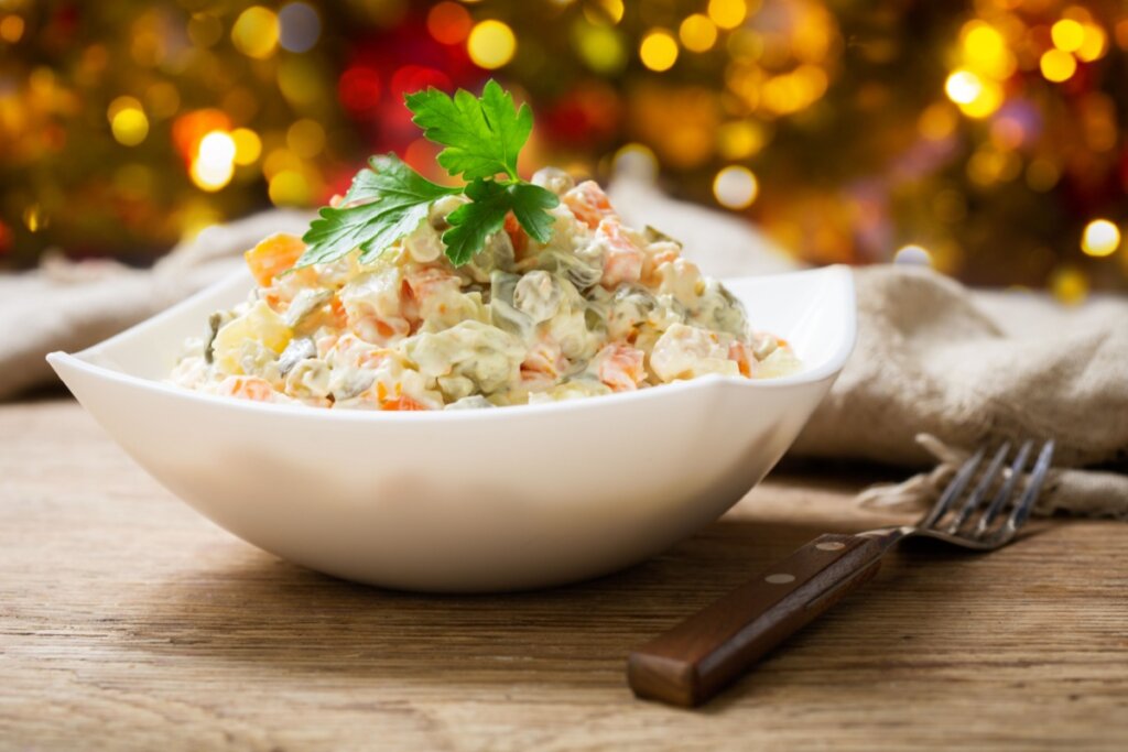 Salada de maionese servida em tigela branca em cima de mesa de madeira com decoração de Natal ao fundo