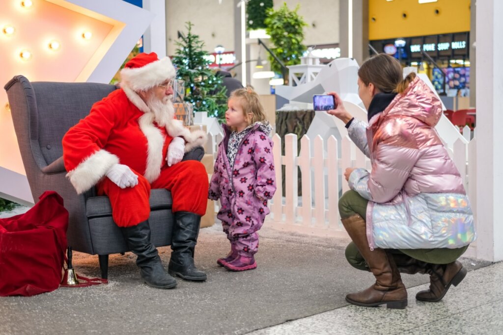 Papai Noel sentado em uma poltrona conversando com uma criança em pé enquanto a mulher ao lado registra a cena com o celular em shopping