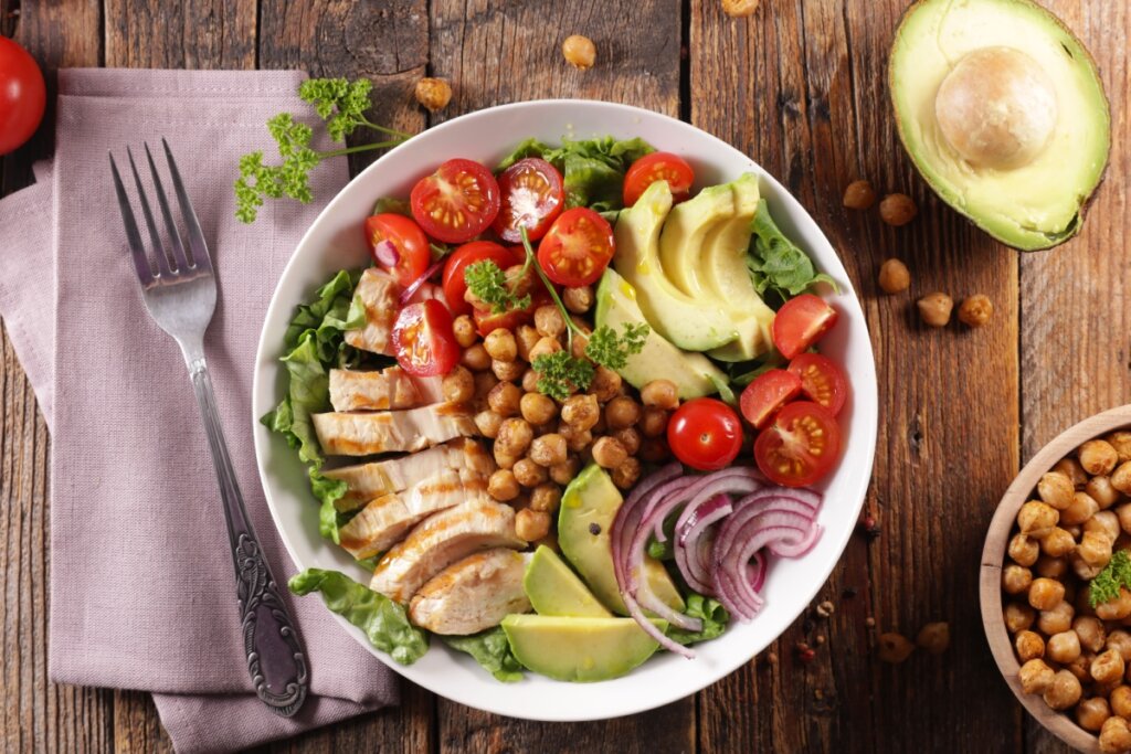 Buddha bowl de frango, grão-de-bico, abacate, tomate, cebola-roxa e alface, ao lado de garfo, lenço, abacate cortado e grão-de-bico