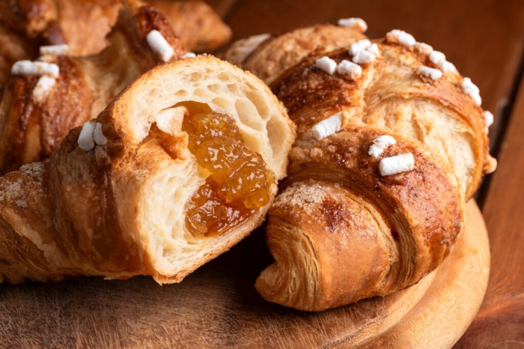 Close-up de um croissant partido ao meio, revelando um interior bem folhado e preenchido com geleia de damasco. A superfície externa está polvilhada com açúcar e decorada com pedaços de açúcar granulado branco. O croissant repousa sobre uma tábua de madeira rústica em tons quentes