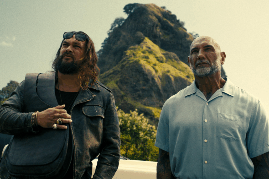 Foto de meio corpo de dois homens em frente a uma colina rochosa e verde sob um céu claro. À esquerda (Jason Momoa), um homem com cabelos longos ondulados, barba e óculos escuros na cabeça usa uma jaqueta de couro e segura uma bolsa a tiracolo. À direita (Dave Bautista), um homem careca com barba grisalha veste uma camisa azul clara de botões. Ambos olham para cima e para o lado com expressões sérias.