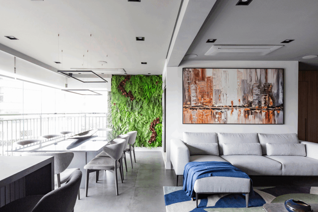 Amplo ambiente integrado de sala de estar e jantar. O destaque é um jardim vertical exuberante que cobre uma parede inteira ao fundo, composto por diversas espécies de samambaias e folhagens verdes com detalhes em tons de roxo. O espaço conta com móveis contemporâneos em tons de cinza, um quadro abstrato grande na parede e luminárias pendentes lineares sobre a mesa de jantar.