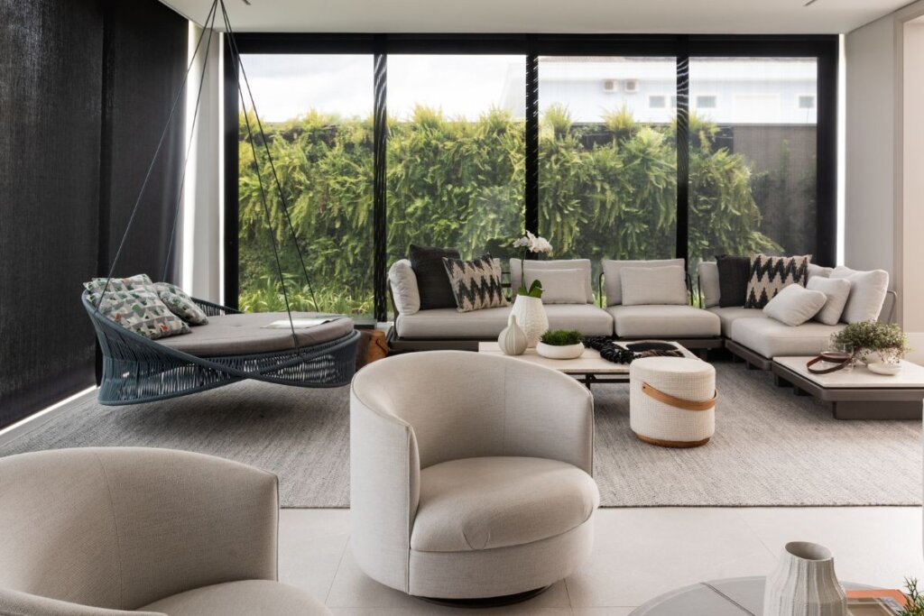 Sala de estar com sofá bege na frente de uma janela preta grande com folhas. Na frente, mesa de centro e poltronas brancas 