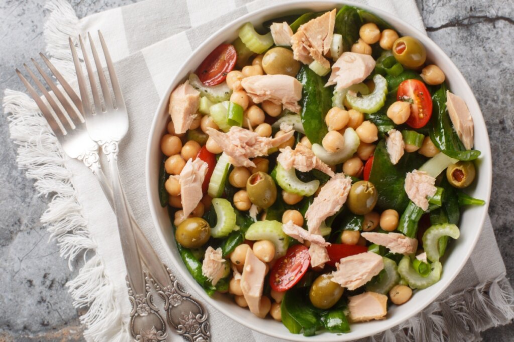 Salada de grão-de-bico com atum, tomate-cereja, espinafre, salsão e azeitonas servida em tigela branca sobre tecido claro