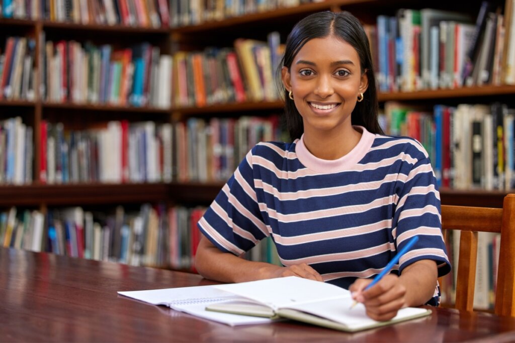 Mulher em ambiente repleto de livros segurando caneta prestes a escrever em caderno