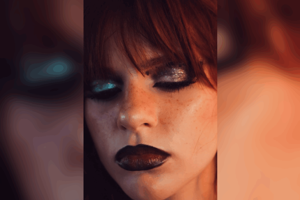 Close de maquiagem gótica com olhos fechados, mostrando sombra preta com partículas de brilho, sardas evidentes no rosto e batom escuro bem definido nos lábios