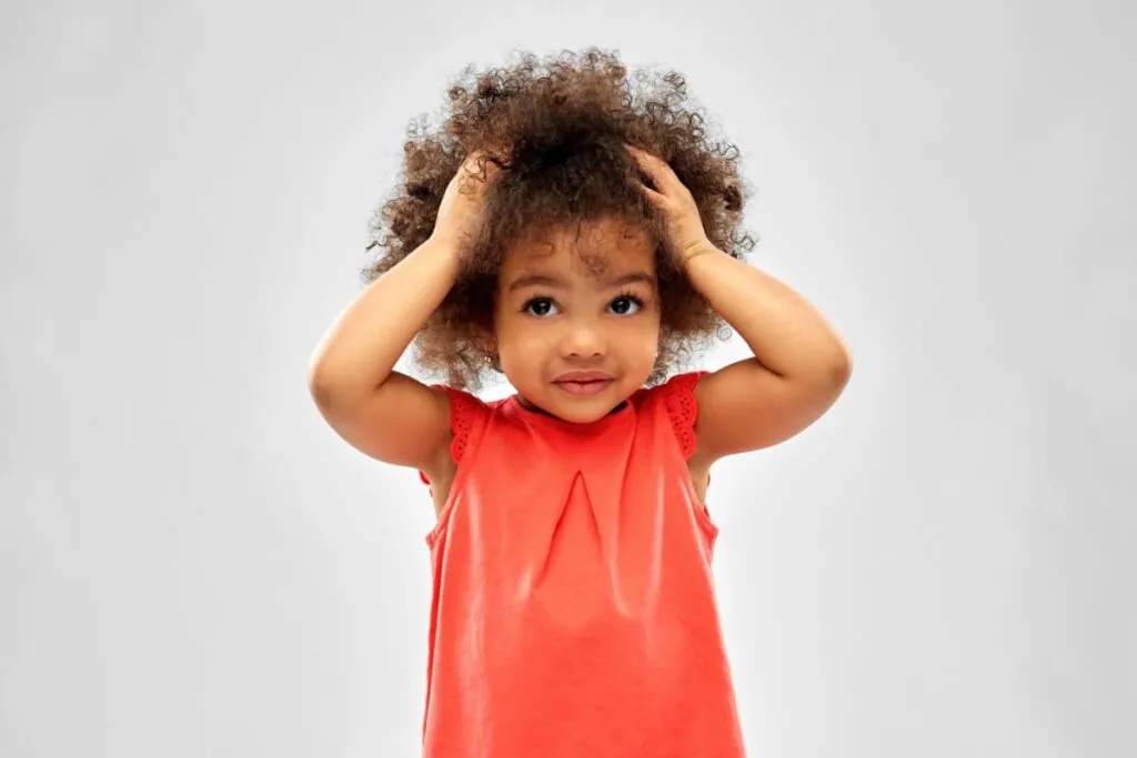 Uma menina negra pequena, com cabelo crespo castanho, posa contra um fundo cinza claro e uniforme. Ela veste uma blusa de cor coral (tom alaranjado) sem mangas, com um detalhe de prega no decote. A criança olha diretamente para a câmera com uma expressão serena e curiosa, enquanto mantém ambas as mãos apoiadas no topo da cabeça, com os cotovelos voltados para fora.