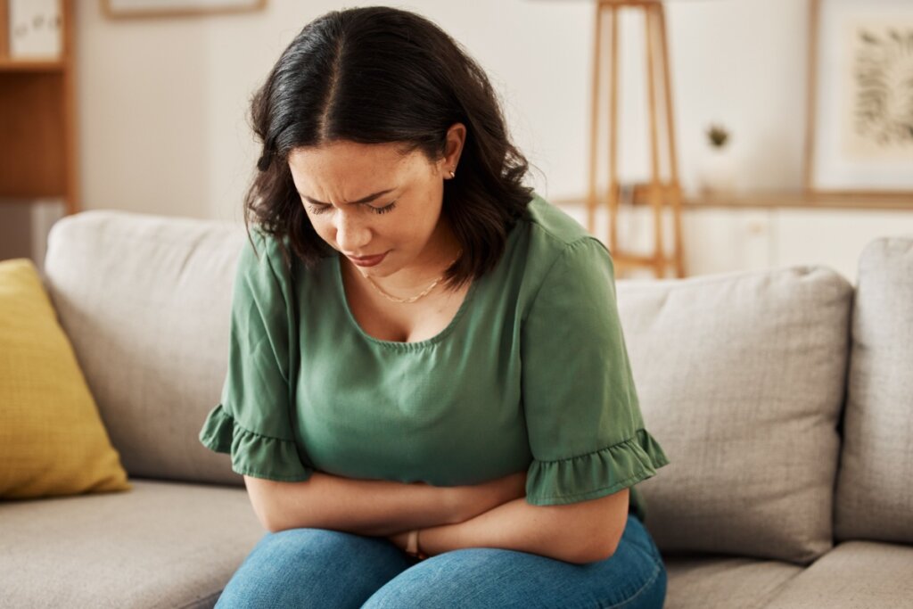 Uma mulher de pele parda está sentada em um sofá cinza, curvada para frente com uma expressão de dor e desconforto. Ela veste uma blusa verde de mangas curtas com babados e calça jeans azul. Seus olhos estão cerrados e ela pressiona as duas mãos contra o abdômen, sugerindo que sente cólica ou dor de estômago. O ambiente ao fundo é uma sala de estar iluminada e decorada em tons neutros.
