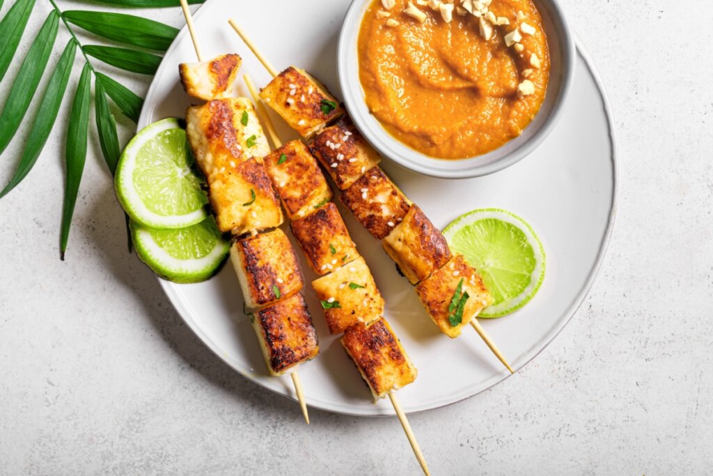 Espetinhos de tofu acompanhados de molho cremoso de amendoim e fatias de limão em prato branco