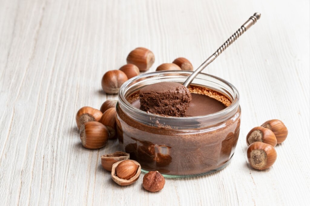 Creme de avelã servido em tigela de vidro com colher, em cima de mesa de madeira com avelãs ao redor