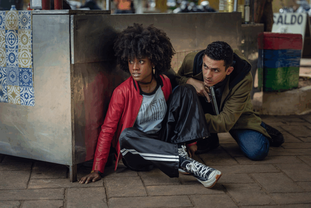 Jovem com cabelo cacheado curto, solto, usando tênis preto, calça jeans, camiseta cinza e preta, jaqueta vermelha sentada no chão ao lado de homem com cabelo curto usando casaco verde e preto, calça jeans e com arma na mão.