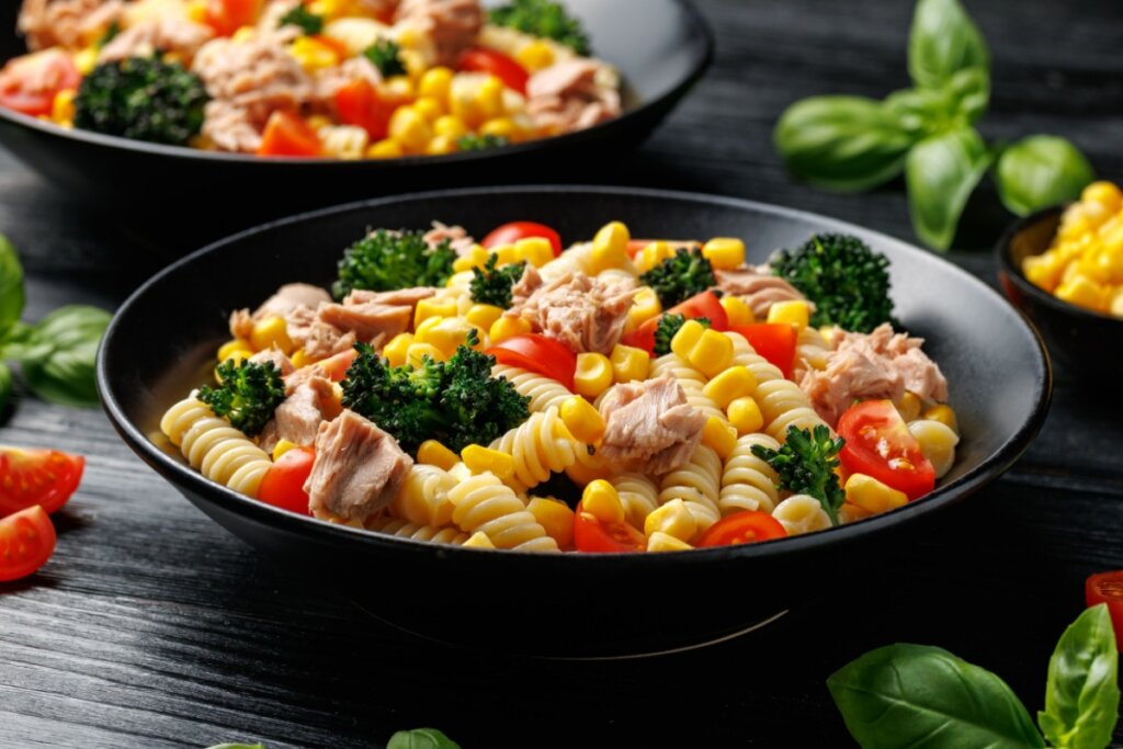 Salada de macarrão com atum, brócolis e milho servido em prato fundo preto em cima de bancada de pedra