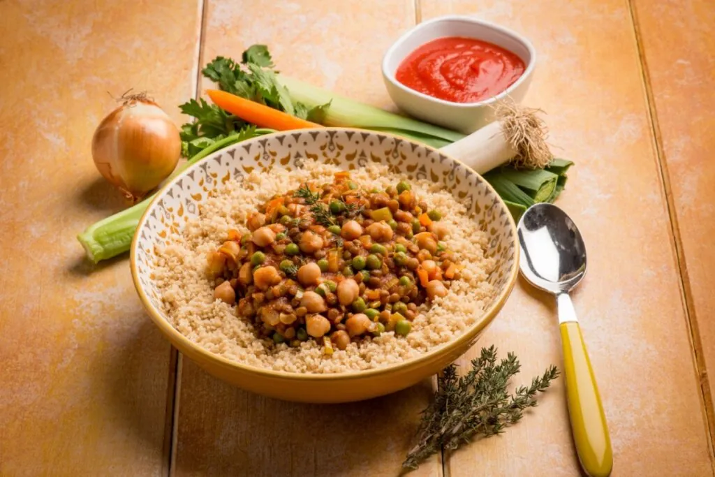 Prato fundo de cuscuz com lentilha, grão-de-bico, ervilha e legumes ao molho servido no centro, acompanhado de ingredientes frescos ao redor