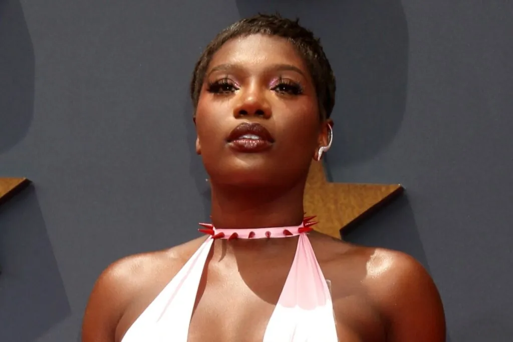 Doechii, usando roupa rosa e com cabelo curto, posando para foto em um fundo cinza 