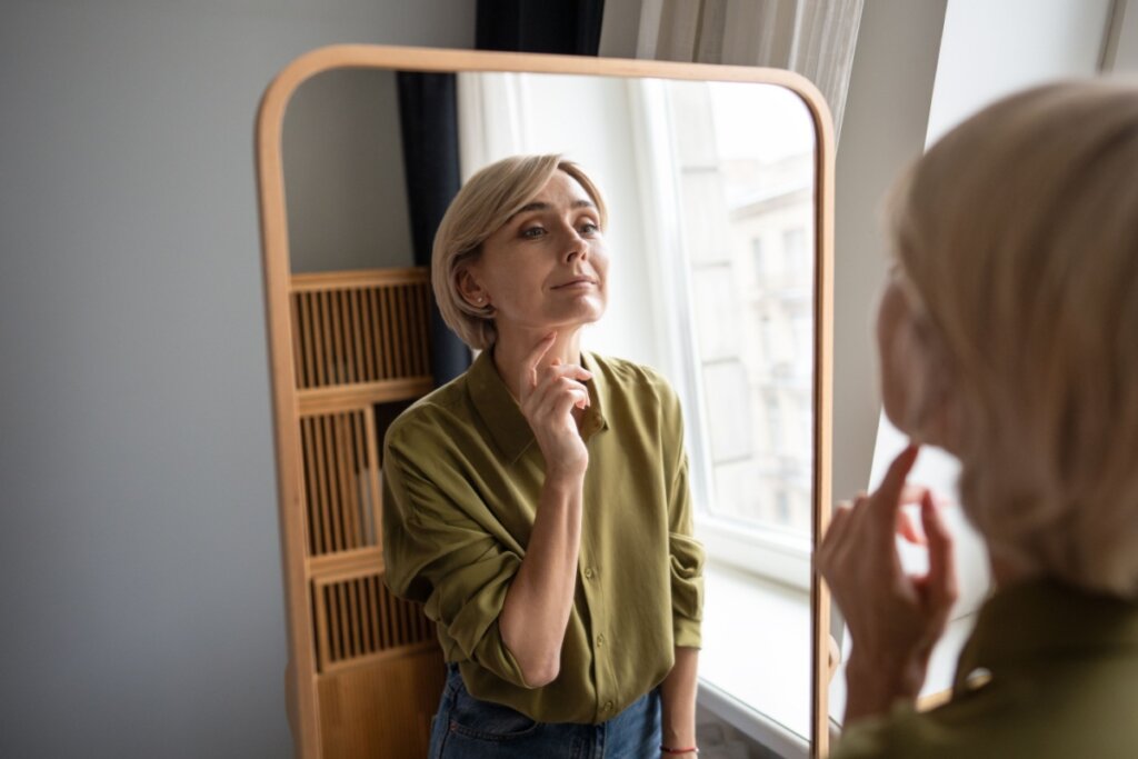 Uma mulher loira de meia-idade, vestindo uma camisa de seda verde oliva e calça jeans, olha para o seu reflexo em um espelho de moldura de madeira clara com cantos arredondados. Ela toca suavemente o queixo com os dedos enquanto se observa. 