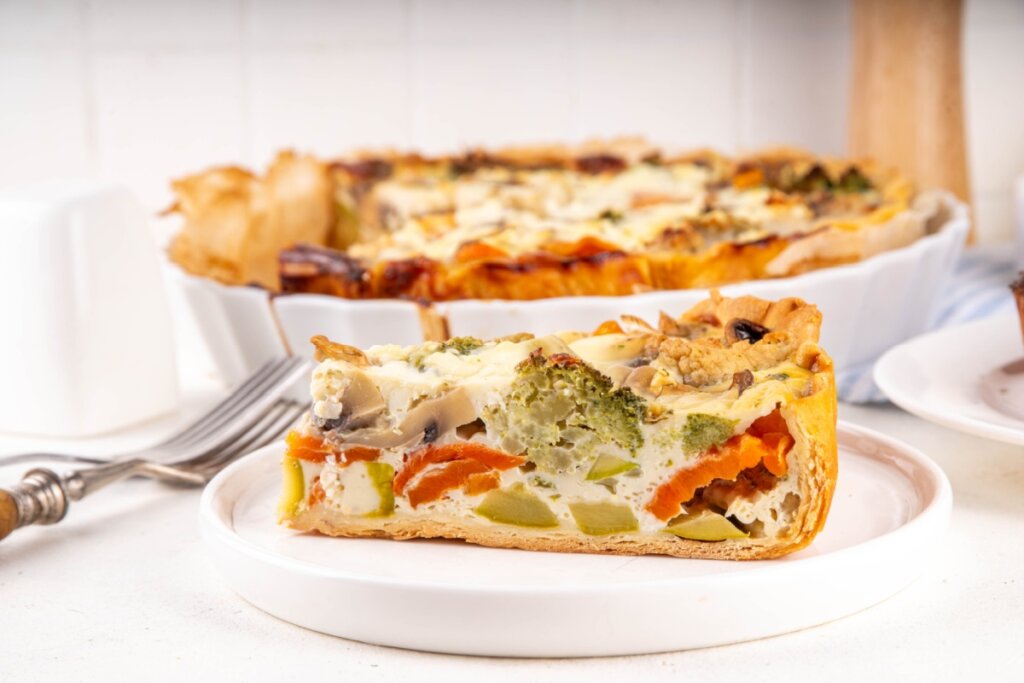 Fatia de torta tipo quiche vegetariana com legumes e cogumelos em prato branco, com forma ao fundo