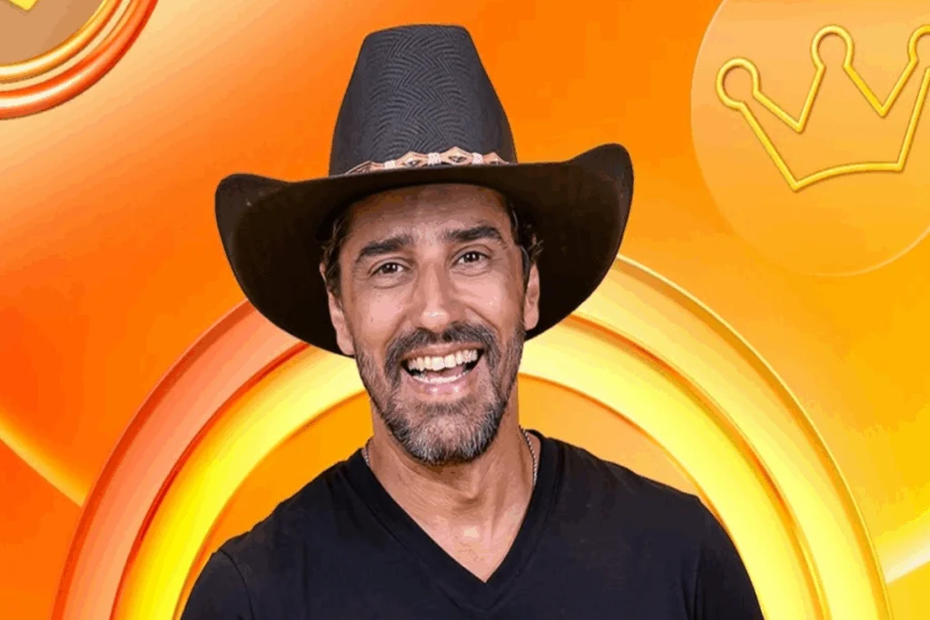 Homem, com cabelo curto e barba, usando chapéu de cowboy preto e camiseta preta em fundo com tons de laranja
