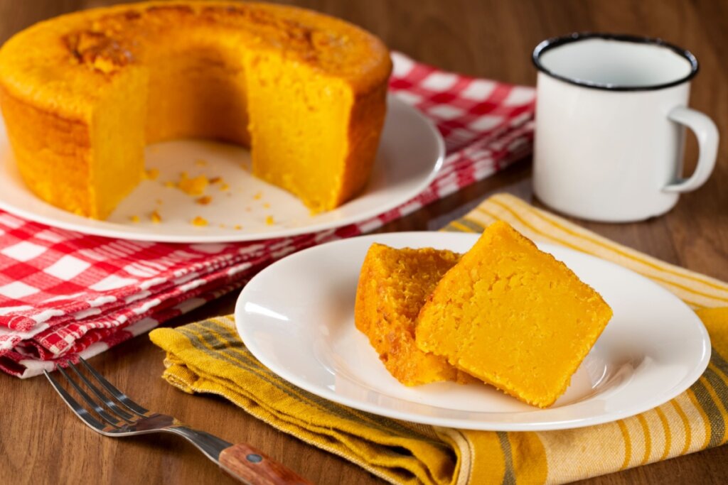 A imagem mostra um bolo de milho caseiro, com textura macia e cor amarela intensa. Um pedaço já foi cortado e servido em um prato, enquanto o restante do bolo aparece ao fundo. Ao lado, há uma xícara, sugerindo que o bolo está sendo servido com café