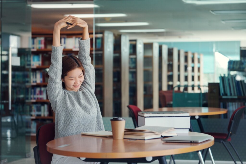 menina de blusa cinza sentada em cadeira de biblioteca e alongando braços para cima. mesa com copo de café e livros