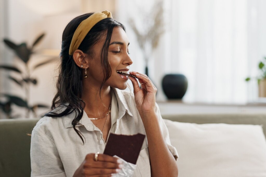 Jovem com o cabelo solto parcialmente usando faixa de cabelo bege, usando camisa cinza e comendo chocolate de olhos fechados