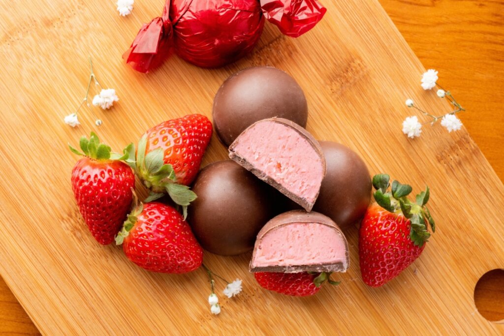 Trufas de chocolate com recheio de morango cortadas ao meio e inteiras, acompanhadas de morangos frescos sobre uma tábua de madeira
