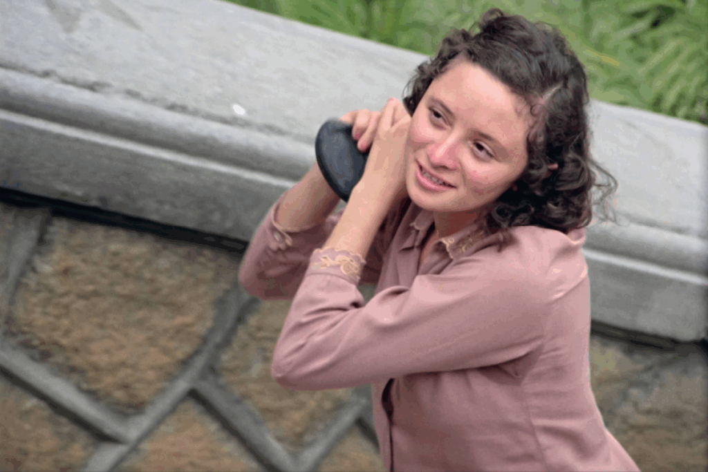 Jovem de cabelos cacheados e blusa rosa sorrindo enquanto segura um objeto escuro próximo ao rosto, em cena ao ar livre