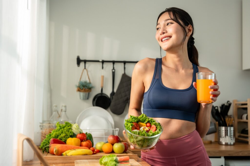 Uma mulher jovem com fisionomia asiática sorri e olha para o lado enquanto segura uma tigela de vidro com salada fresca e um copo de suco de laranja. Ela veste roupas esportivas (top azul e calça rosa) e está em uma cozinha bem iluminada pela luz natural que entra por uma janela. À sua frente, sobre a bancada de madeira, há uma grande variedade de frutas e legumes coloridos, como tomates, pimentões, cenouras, maçãs e bananas. O ambiente transmite uma atmosfera de bem-estar e alimentação saudável.