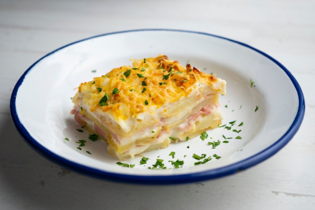 Uma fatia quadrada de lasanha de batata é servida no centro de um prato raso de esmalte branco com borda azul. A lasanha apresenta camadas visíveis de fatias finas de batata clara, intercaladas com molho branco (bechamel) cremoso e fatias de presunto rosado. O topo está coberto por uma camada generosa de queijo gratinado e dourado.