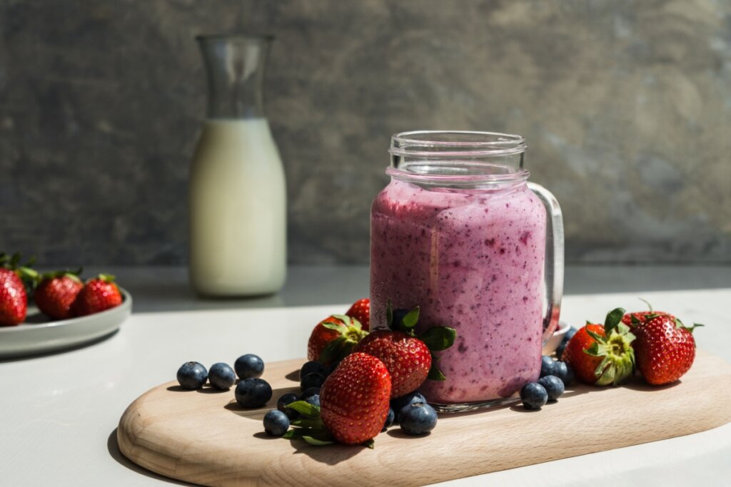 A imagem mostra uma vitamina cremosa de frutas vermelhas servida em um copo de vidro com alça. A bebida tem coloração rosada, indicando a mistura de ingredientes como morango e mirtilo.
Ao redor, aparecem frutas frescas — morangos e blueberries — além de uma jarra de leite ao fundo
