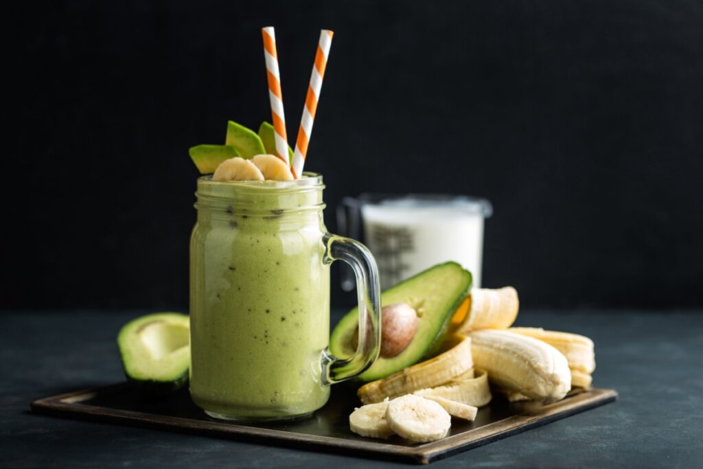 Vitamina proteica de abacate com banana servido em copo de vidro com canudinho branco com listras laranja e abacate cortado ao meio e banana em rodelas ao lado em fundo preto
