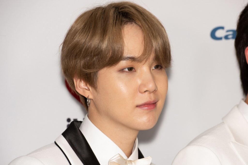 Homem com cabelo castanho claro partido ao meio com franja, usando camisa branca e terno bege com detalhes em preto e gravata borboleta bege