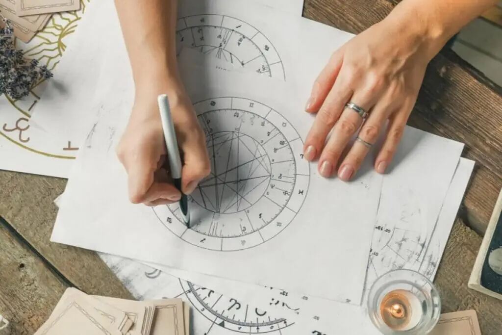Uma visão aérea em close-up mostra as mãos de uma pessoa trabalhando sobre uma mesa de madeira rústica com mapas de mapa astral impressos em papel branco. A mão esquerda segura uma caneta branca e preta, apontando para as marcações de um grande gráfico circular astrológico repleto de linhas entrecruzadas e símbolos do zodíaco. A mão direita, usando dois anéis prateados, repousa aberta sobre o papel. Ao redor, nas bordas da mesa, veem-se cartas de Tarot empilhadas, ramos de lavanda seca e uma pequena vela acesa dentro de um suporte de vidro.