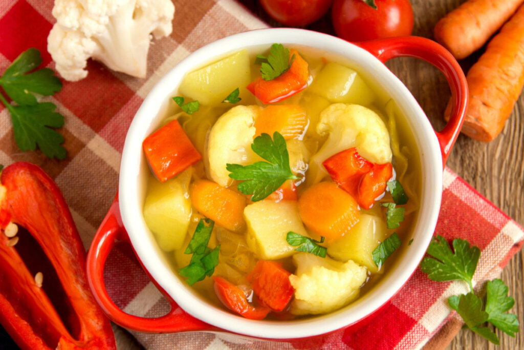Panela vermelha com sopa de cenoura e batata com coentro em cima de uma mesa de madeira com vegetais ao redor 