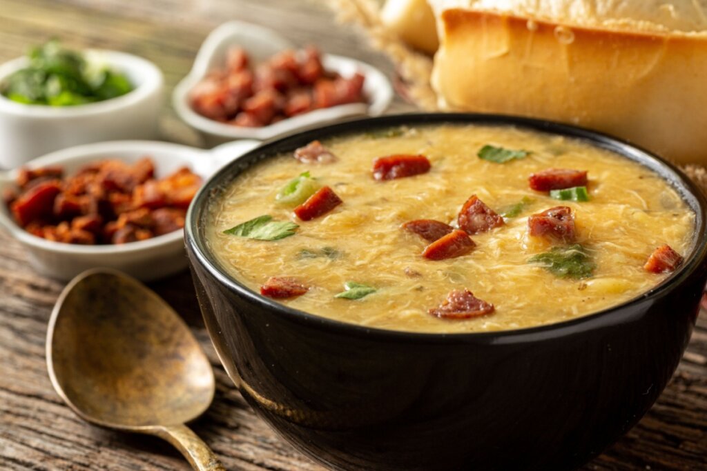 Uma tigela preta cheia de caldo de frango cremoso e amarelado, salpicado com pedaços picados de linguiça calabresa frita, rodelas de cebolinha e folhas de salsa. A tigela está posicionada sobre uma mesa de madeira rústica. À esquerda, em primeiro plano, vê-se uma colher de metal envelhecido e, ao fundo, pequenos recipientes brancos com acompanhamentos (como linguiça, bacon e cheiro-verde), além de um pedaço de pão à direita.