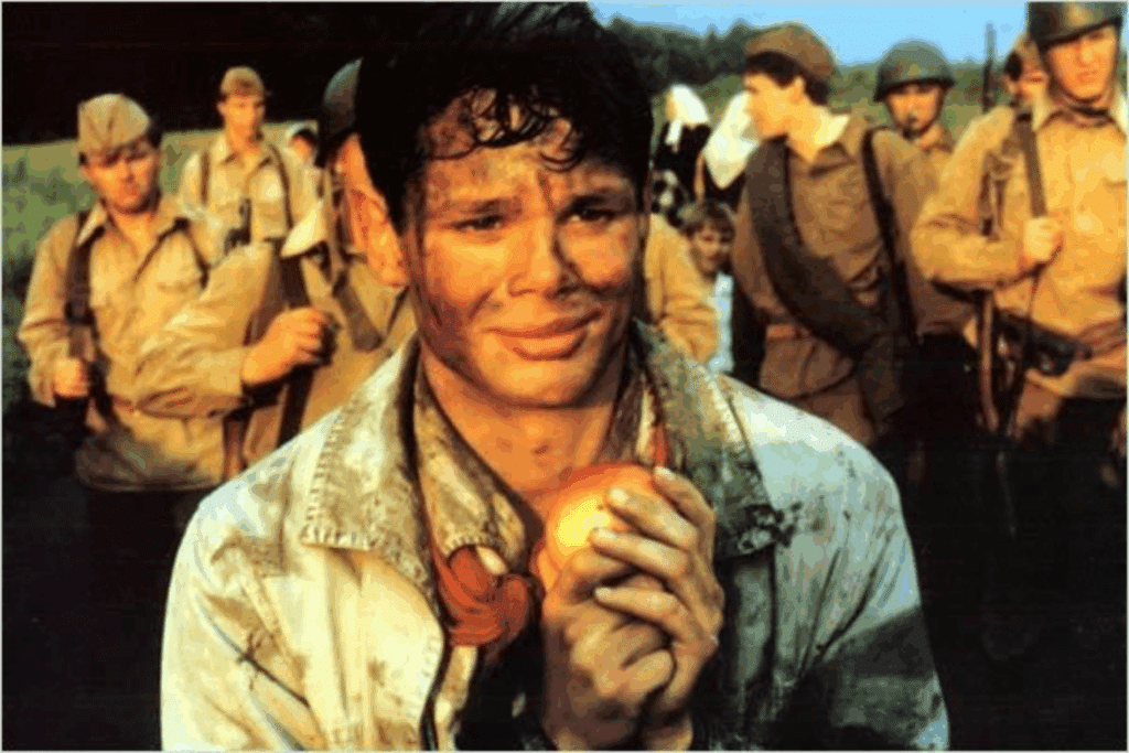 Em primeiro plano, um jovem soldado com o rosto sujo de terra sorri de forma contida enquanto segura uma maçã vermelha com as duas mãos, perto do peito. Ele veste uma jaqueta clara manchada e um lenço vermelho no pescoço. Ao fundo, vários outros soldados uniformizados caminham em um campo aberto sob a luz do dia.
