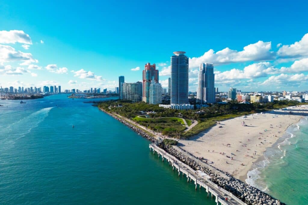 Uma vista aérea de Miami Beach, mostrando a longa faixa de areia com banhistas, a entrada do porto e uma fileira de arranha-céus à beira-mar.
