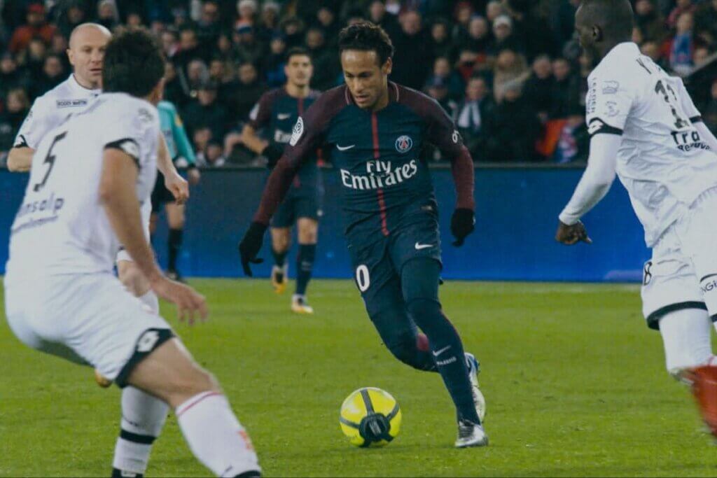 Uma imagem de corpo inteiro de Neymar Jr., um dos melhores jogadores de futebol da atualidade, com a camisa azul e vermelha do Paris Saint-Germain, o uniforme número dez. Ele está driblando com a bola, com dois jogadores adversários ao seu redor. O fundo é um campo de futebol.
