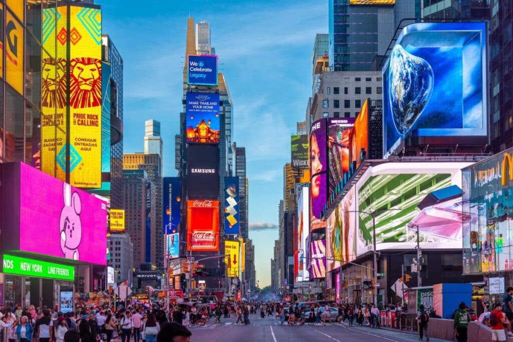 Uma foto vibrante de nível de rua de Times Square, Nova York, com inúmeros outdoors luminosos gigantes cobrindo arranha-céus e uma multidão de pedestres.