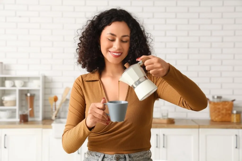 mulher de blusa marrom sorrindo, segurando bule e despejando café em caneca cinza
