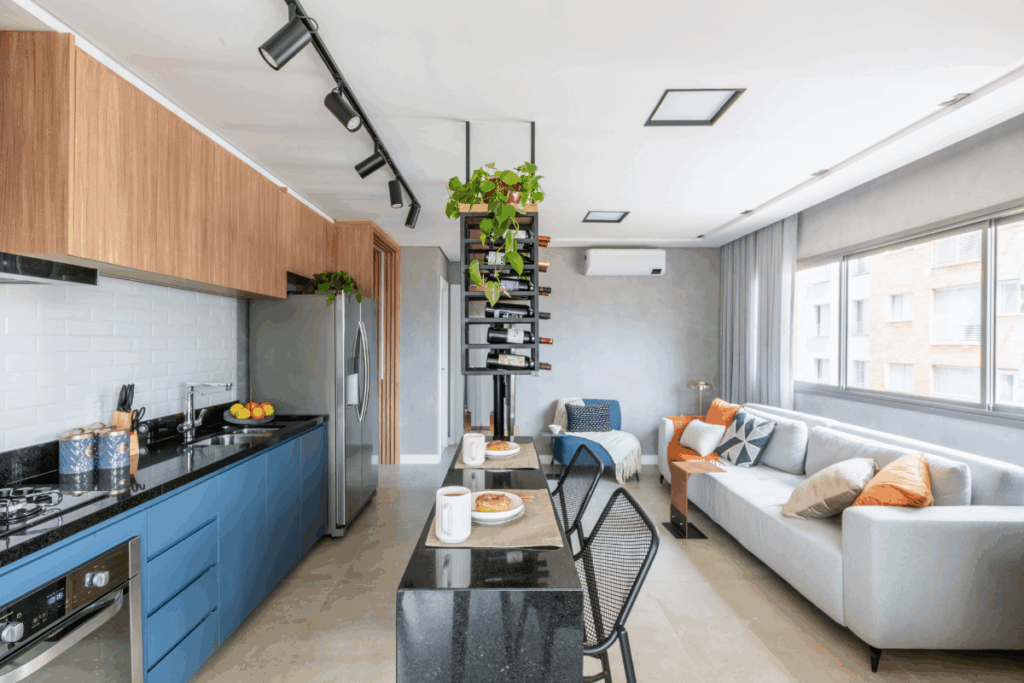 Apartamento de conceito aberto com cozinha integrada à sala de estar. À esquerda, a cozinha possui armários inferiores azuis, bancada de granito preto, revestimento de azulejos brancos e armários superiores de madeira. No centro, uma bancada preta com banquetas serve de divisória e mesa de refeições, sob uma adega vertical suspensa com plantas. À direita, a sala de estar conta com um sofá cinza claro decorado com almofadas laranjas e estampadas, posicionado em frente a uma grande janela.