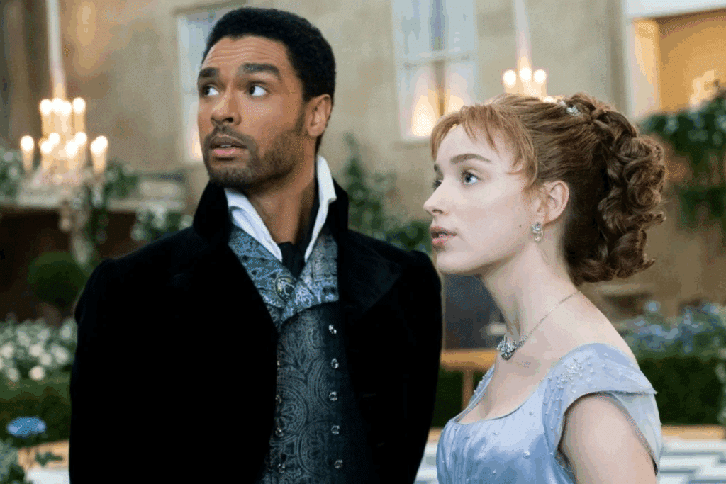 Um homem negro vestido em trajes de gala pretos e um colete estampado olha para cima surpreso, ao lado de uma mulher branca de cabelos presos em um penteado clássico e vestido lilás, que também olha na mesma direção.
